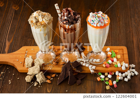 Three sweet milkshakes with chocolate, marshmallow, halva and whipped cream at a wooden board on table background. Three sweet milkshakes with chocolate, marshmallow, halva and whipped cream at a wooden board on table background. 122604001