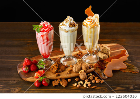 Three sweet milkshakes with nuts, caramel, strawberry and whipped cream at a wooden board on table background. 122604010
