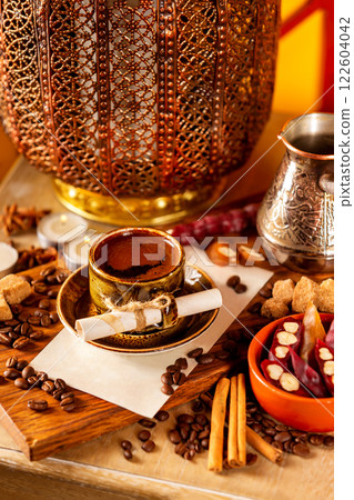Cup of turkish black coffee in a plate with blank paper sheet on a wooden board at decorated table with candles, beans and sweets background. Cup of turkish black coffee in a plate with blank paper sheet on a wooden board at decorated table with candles, beans and sweets background. 122604042