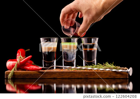 Three alcohol shooter layered cocktails on a wooden board served with barman spoon and chili pepper isolated at black background. Barman is making hiroshima cocktail. Three alcohol shooter layered cocktails on a wooden board served with barman spoon and chili pepper isolated at black background. Barman is making hiroshima cocktail. 122604208