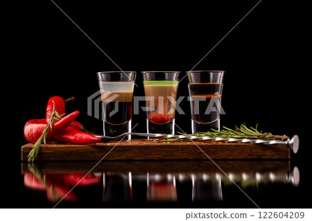 Three alcohol shooter layered cocktails on a wooden board served with barman spoon and chili pepper isolated at black background. 122604209