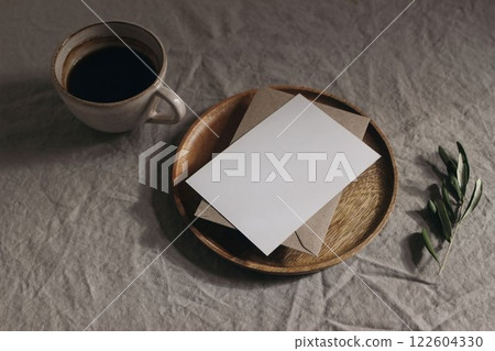 Overhead view of blank greeting card invitation mockup, envelope on wooden plate. Cup of coffee olive branch. Linen table cloth in sunlight. Moody summer breakfast still life stationery. Working space Overhead view of blank greeting card invitation mockup, envelope on wooden plate. Cup of coffee olive branch. Linen table cloth in sunlight. Moody summer breakfast still life stationery. Working space 122604330