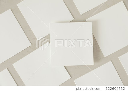 Many empty white square business card greeting card mockups with soft shadows lying diagonally on beige textured wooden table background. One card above. Flat lay, top view. Stationery composition. 122604332