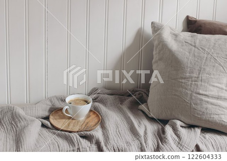 Scandinavian living room, bedroom. Ceramic mug of hot coffee. Linen throw blanket, velvet cushions pillows. Breakfast in bed. Soft sunlight. Warm neutral tones. White wooden beadboard wall background. 122604333