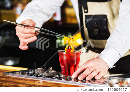 Bartender is holding orange rind in tongs decorating red cocktail at bar background. Bartender is holding orange rind in tongs decorating red cocktail at bar background. 122604657