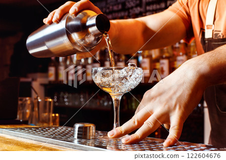 Male bartender is making cocktail pouring alchohol from shaker to glass at bar background. 122604676