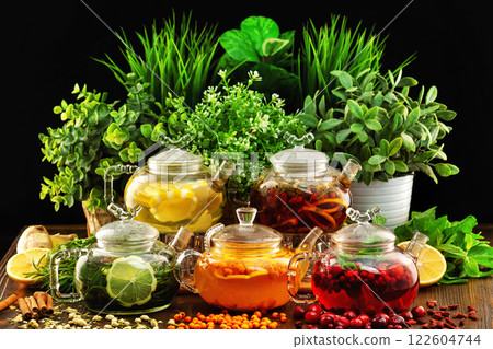 Set of colorful glass tea pots decorated with green plants and berries on a wooden table board at dark background. 122604744