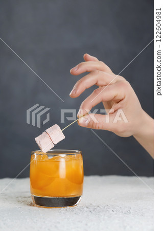 Bartender's hand is holding stick with marshmallow over orange cocktail at grey wall background. 122604981