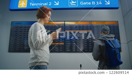 Airport Terminal: Woman with Luggage and Ticket Checks Her Flight on Digital Departure Board 122609514