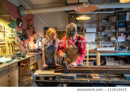 Beginner woman watch master carpenter operate saw machine in workshop. Husband and wife collaborate Beginner woman watch master carpenter operate saw machine in workshop. Husband and wife collaborate 122610425