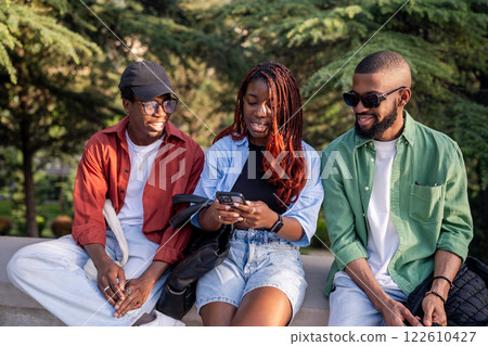 Lively African American friend sit in park, phone in hand. Woman shares trending social media post Lively African American friend sit in park, phone in hand. Woman shares trending social media post 122610427