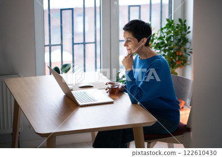Content interested mature woman working on laptop, sipping tea and watching screen. Reading articles 122610430