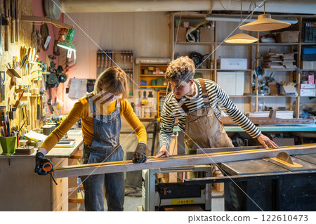 Woman and man carpenters work together on circular saw with wooden beam in workshop. Mentorship 122610473