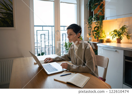 Tired focused woman works at laptop, take notes in notebook at home. Balances remote work and study 122610474