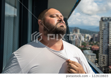 Plump man wave shirt collar to cool down on balcony, dealing with intense heat. Discomfort, sweating Plump man wave shirt collar to cool down on balcony, dealing with intense heat. Discomfort, sweating 122610603