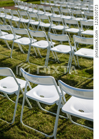 Folding chairs standing in row on green grass, casting shadow on ground. Outdoor party preparation 122610723