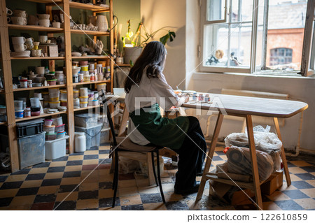 Focused woman artisan painting ceramic ware in workshop. Detailed work with clay dishes. Rearview Focused woman artisan painting ceramic ware in workshop. Detailed work with clay dishes. Rearview 122610859