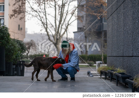 Kind, warm-hearted stylish guy in trendy outfit sitting on haunches, caressing hungry street dog 122610907