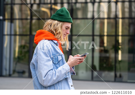 Pleased hipster guy standing outside with smartphone, looking screen typing message in social media. Pleased hipster guy standing outside with smartphone, looking screen typing message in social media. 122611010