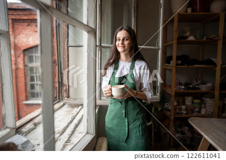 Smiling woman potter drinks tea from handmade ceramic mug by window in workshop, enjoying creation 122611041