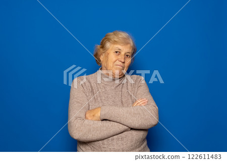 Caucasian woman about 70s wearing dark beige sweater shrugging shoulders, feeling confused and unsure, doubting with arms crossed. Isolated over blue background Caucasian woman about 70s wearing dark beige sweater shrugging shoulders, feeling confused and unsure, doubting with arms crossed. Isolated over blue background 122611483