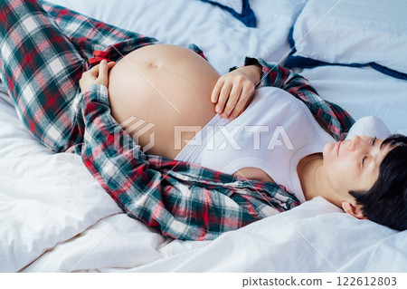 Pregnant woman lying on bed, touching her belly, showcasing joy and anticipation. Expecting mother relaxing in pajama during last trimester of pregnancy. Motherhood, preparation and expectation. Pregnant woman lying on bed, touching her belly, showcasing joy and anticipation. Expecting mother relaxing in pajama during last trimester of pregnancy. Motherhood, preparation and expectation. 122612803