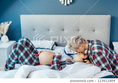 Mixed race couple are waiting for a baby. Husband and his pregnant wife in cozy pajamas relaxing on the bed at modern home bedroom. Romantic moments for pregnant couple, warm atmosphere of love. Mixed race couple are waiting for a baby. Husband and his pregnant wife in cozy pajamas relaxing on the bed at modern home bedroom. Romantic moments for pregnant couple, warm atmosphere of love. 122612804