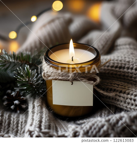 Scented candle in glass jar on soft grey jersey fabric, Winter product photo. Warm glow of scented candle creates cozy winter atmosphere. Scented candle jar with blank label mockup 122613083