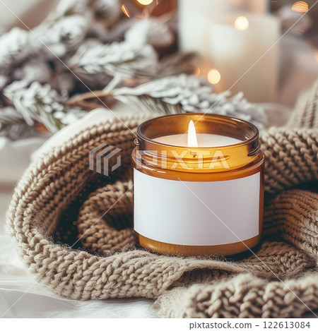 Scented candle in glass jar on soft grey jersey fabric, Winter product photo. Warm glow of scented candle creates cozy winter atmosphere. Scented candle jar with blank label mockup 122613084