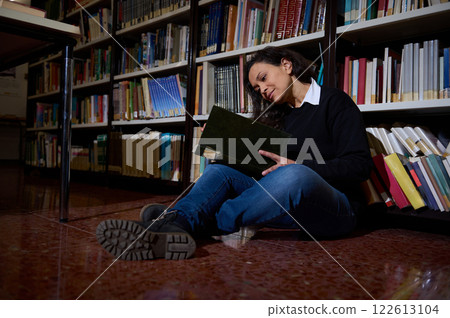 Young woman reading a book while sitting on the library floor Young woman reading a book while sitting on the library floor 122613104