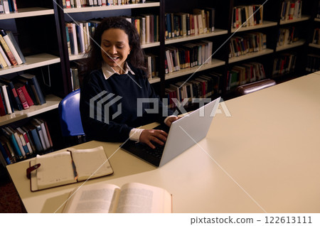Smiling woman using a laptop in a library for studying or working Smiling woman using a laptop in a library for studying or working 122613111