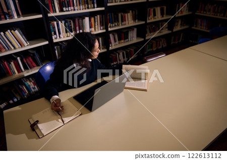 Individual studying in a library using books and a laptop at a table Individual studying in a library using books and a laptop at a table 122613112
