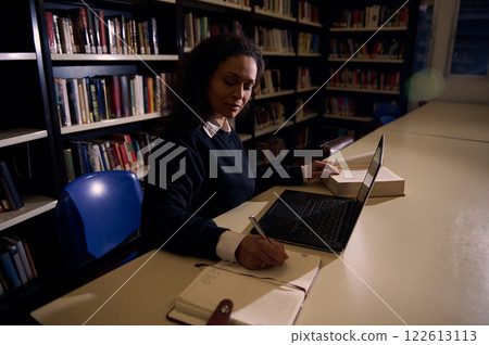 Woman studying in library with a laptop and open books around her Woman studying in library with a laptop and open books around her 122613113
