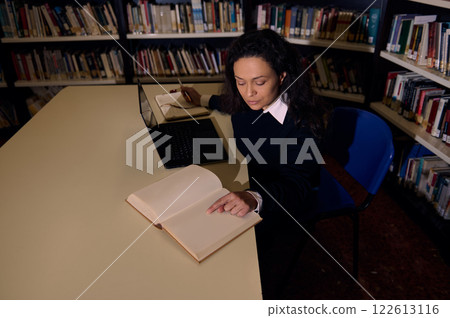 Woman reading in a library while studying with a laptop and notebook 122613116