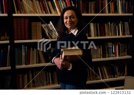 Smiling Woman Holding Books in a Library Surrounded by Shelves Smiling Woman Holding Books in a Library Surrounded by Shelves 122613132