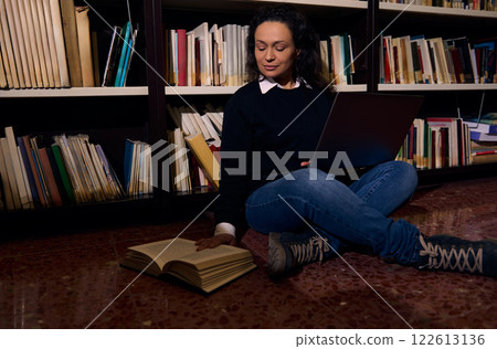Woman studying with a laptop and book in a library setting Woman studying with a laptop and book in a library setting 122613136