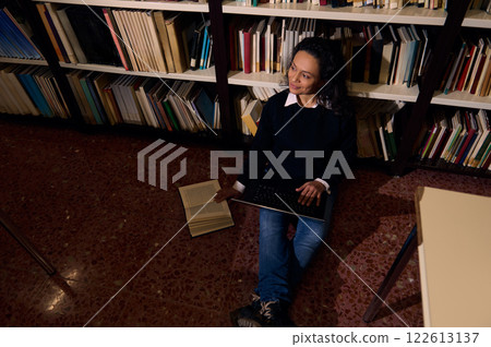 Woman studying with a laptop and book in a library for knowledge Woman studying with a laptop and book in a library for knowledge 122613137