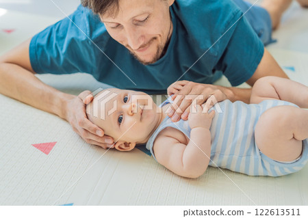 A baby lies on a soft children play mat while their father hands gently support and protect them. Heartwarming moment of care and connection. Parenthood, safety, and love concept 122613511