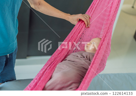 A 3-month-old baby peacefully falls asleep in a soft muslin swaddle while cradled in a cozy hammock. A serene and tender moment of comfort and relaxation. Baby care, rest, and tranquility concept 122614133