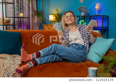 Successful rich smiling woman holding smartphone waving dollar bill money fan sitting on home sofa 122614134