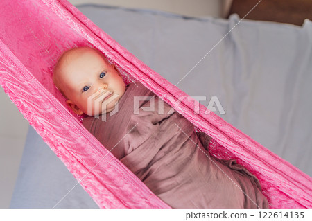 A 3-month-old baby peacefully falls asleep in a soft muslin swaddle while cradled in a cozy hammock. A serene and tender moment of comfort and relaxation. Baby care, rest, and tranquility concept 122614135