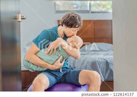 A father sits on a fitness ball in a cozy bedroom, gently rocking his baby wrapped in a soft muslin swaddle. A nurturing and soothing moment of fatherhood and care. Parenting, bonding, and comfort A father sits on a fitness ball in a cozy bedroom, gently rocking his baby wrapped in a soft muslin swaddle. A nurturing and soothing moment of fatherhood and care. Parenting, bonding, and comfort 122614137