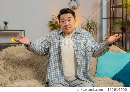 Confused Asian handsome man feeling embarrassed about ambiguous question sitting on sofa at home Confused Asian handsome man feeling embarrassed about ambiguous question sitting on sofa at home 122614700