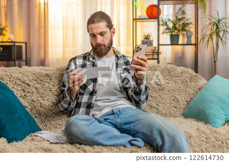 Young man sorting bills at home, sofa budget check, phone in hand, multiple papers, calm focus vibe Young man sorting bills at home, sofa budget check, phone in hand, multiple papers, calm focus vibe 122614730