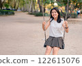 Young Girl Enjoying Sunny Day on Swing in Park Surrounded by Green Trees and Pathway 122615076