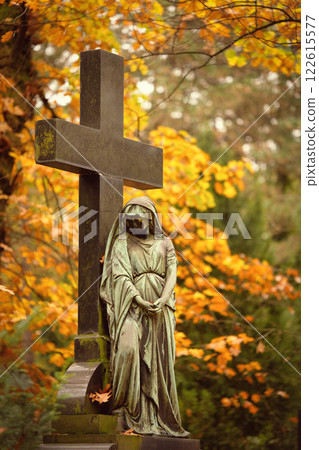 a female cemetery statue with her face black from weathering leans with folded hands against a cross in front of glowing autumn leaves in the background a female cemetery statue with her face black from weathering leans with folded hands against a cross in front of glowing autumn leaves in the background 122615577