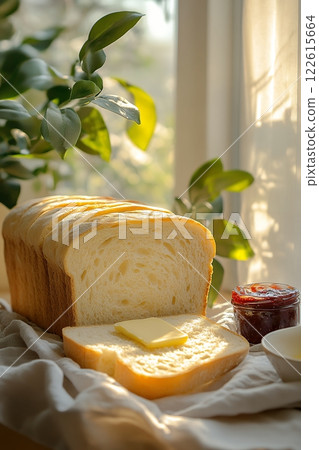 Sunlight streams through a window onto a loaf of sliced sourdough bread, topped with butter. A jar of jam and a plant complete the inviting scene. Perfect for a cozy breakfast! 122615664