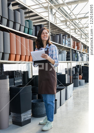 Woman worker in garden center wearing uniform, surrounded many pots for plants on racks and shelves Woman worker in garden center wearing uniform, surrounded many pots for plants on racks and shelves 122616095