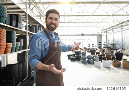 Man worker in garden center wearing uniform, arranging many pots for plants on racks and shelves 122616105