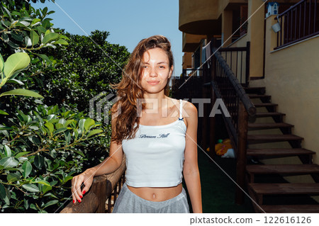 A cheerful young woman poses confidently outdoors by a wooden staircase, surrounded by lush greenery A cheerful young woman poses confidently outdoors by a wooden staircase, surrounded by lush greenery 122616126
